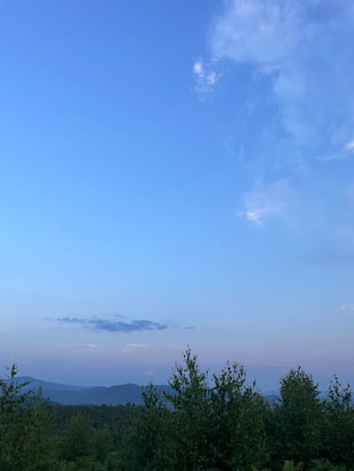 The image depicts a serene landscape scene from Rokosovo, Ukraine. The vast, clear blue sky dominates the upper portion of the frame, with a few wispy white clouds scattered across it. As the eye moves towards the horizon, the sky transitions into softer hues of pale blue and pink, suggesting it is either early morning or late evening.
On the right side of the image, a lush green forest lines the foreground. The foliage is dense and appears healthy, with the leaves of trees catching the soft light. Beyond the forest, a series of rolling hills or mountains recede into the distance, their forms gradually becoming more muted and blending with the hazy sky. This layering of distant peaks creates a sense of depth and expanse within the scene.
The overall atmosphere is peaceful and tranquil. There are no people or specific objects to identify, focusing the viewer's attention on the natural beauty of the landscape. The soft lighting and gentle colors evoke a sense of calm and stillness. The time of day is likely during the twilight hours, given the sky's coloration. The location is identifiable as Rokosovo, Ukraine, a region known for its natural landscapes. There is no visible text in the image.