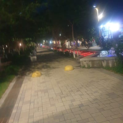 The image depicts a nighttime scene in what appears to be a public space or park in Turkistan, Kazakhstan. The scene is illuminated by artificial lights. A paved walkway with a geometric pattern dominates the foreground and extends into the middle ground. Along the left side of the walkway, there is a raised platform or ledge with red lighting. People are gathered on the platform. Trees and greenery are also visible. Two yellow hemispherical bollards are present on the walkway. In the background, more lights and possibly buildings are seen. The overall environment suggests an evening gathering in a well-lit urban park.