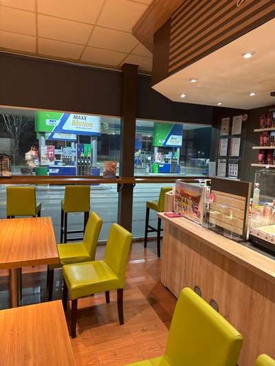The scene depicts the interior of a modern, unoccupied cafe or convenience store located in Gorno Aleksandrovo, Bulgaria, looking out onto an illuminated OMV MAXX Motion gas station at night. The interior features a light-colored wooden laminate floor, square wooden tables with metallic bases, and numerous lime-green upholstered chairs and bar stools. A light-wood service counter on the right side of the frame displays various brochures, including one with a colorful cityscape illustration, and retail items. Behind this counter, a dark grey wall holds framed documents and shelves stocked with products. The ceiling is a white suspended panel system with recessed lighting. Through large glass windows, the exterior OMV gas station is visible with its distinctive green and blue fuel pumps and overhead canopies bearing the "MAXX Motion Performance Fuels" branding. White lines, potentially a pedestrian crossing, are visible on the pavement directly outside the window. One individual is partially discernible outdoors near a fuel pump in the mid-ground. No direct actions or interactions are occurring within the visible cafe space or immediately outside.