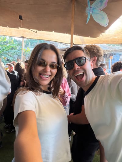 The image captures two individuals in the foreground posing for a selfie at an outdoor gathering in Pardes Hana - Karkur, Israel. The female on the left, with brown hair, wears gold-framed rectangular sunglasses and a light cream short-sleeved t-shirt with a black collar; she is smiling broadly with her tongue extended. Her right arm is visible, extended to hold the camera. The male on the right, with dark hair, wears black-framed sunglasses and a matching light cream short-sleeved t-shirt with a black collar; he is smiling with an open mouth. Both subjects exhibit expressive, joyful facial expressions. In the blurred background, numerous other individuals are visible, standing and engaged in various interactions. The entire scene is covered by a large, light brown canvas canopy, supported by wooden poles, with a light blue and purple butterfly decoration suspended from it on the right. Lush green vegetation is discernible outside the covered area to the left and rear, with additional covered structures further in the background. The ground underfoot appears to be green turf, and the scene is illuminated by strong daylight.
