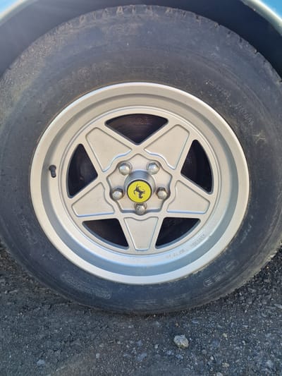 The image presents a close-up view of a car's wheel, featuring a tire mounted on a five-spoke alloy rim. The vehicle is stationary on a surface composed of asphalt and small gravel, under clear daylight conditions. A sliver of a blue car body is visible in the upper left corner.
The tire is black and exhibits signs of age and wear, including fine cracks on the sidewall. Prominently visible text on the tire sidewall includes "RADIAL X" and "TRX," identifying it as a Michelin TRX radial tire. The size specification "240/55VR 415" is clearly readable, denoting a 240mm section width, 55% aspect ratio, V speed rating, and a unique 415mm rim diameter (approximately 16.3 inches). Other partial text includes "TUBEL***S" (likely "TUBELESS"), "MAX LOAD", and "MADE IN FRANCE".
The rim is a light silver or grey color, designed with five spokes forming a star pattern. At its center, a yellow cap displays the iconic black prancing horse logo of Ferrari. The word "Ferrari" is embossed or engraved on one of the spokes, and the rim size "160 TR 415" is also visible, confirming the 415mm diameter matching the tire. Several lug nuts secure the wheel, and a black valve stem is present. The distinct tire and rim specifications, coupled with the Ferrari branding, suggest this is a wheel from a classic Ferrari model, commonly found on vehicles from the late 1970s and 1980s. No individuals or specific activities are depicted. The scene is an outdoor setting, consistent with its provided location context of South Tyneside, United Kingdom.