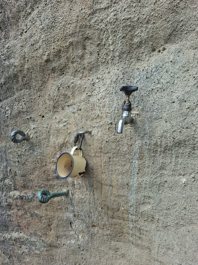 The image depicts a textured concrete wall, likely in Děčín, Czechia, featuring three distinct metal fixtures. On the left, two eye-shaped metal loops are embedded in the wall, one above the other, with the lower one appearing to be made of twisted green metal. To the right of these loops, a light yellow enamel mug with a blue rim is suspended by a metal clip and carabiner from a U-shaped metal bracket. Further to the right and slightly higher on the wall is a silver-colored water tap with a black knob. The wall surface exhibits an uneven, rough texture with visible stains and streaks, suggesting it may be an exterior wall exposed to the elements. No individuals or ongoing actions are visible in the scene.