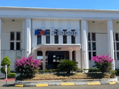 The image shows the front facade of the KAI Cilacap train station, located in Desa Tambakreja, Indonesia. The building is white with large pillars and a prominent signage that reads "KAI Cilacap". In front of the building, there are several windows and an entranceway. The surrounding area is landscaped with green bushes, a palm tree, and a vibrant pink flowering bush. The sky is clear and blue, indicating a sunny day. The area in front of the building appears to be a driveway or parking lot, with yellow and black striped curbs. The overall impression is that of a well-maintained public building in a tropical environment.