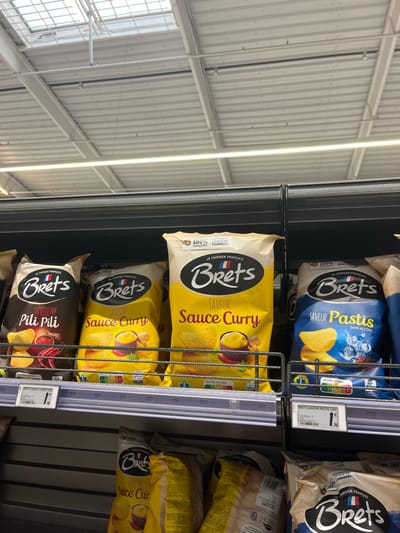 The image shows four bags of Bret's potato chips, on a shelf in a supermarket, located in Saint-Brice-Courcelles, France. Each bag features a different flavor, displayed on the front with a picture of the chips. The first bag features the flavor "Pili Pili", the second bag is "Sauce Curry", the third bag is also "Sauce Curry" and the last bag is "Pastis Sans Alcool". There is a price tag attached to the last bag, indicating a price of 1.70 euros. The background of the image is a white ceiling, with a skylight that appears to be made of glass or plastic. There are no people in the scene.