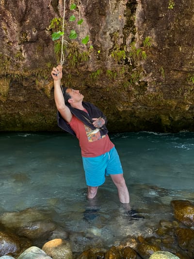 A man stands in shallow, clear turquoise water near a rocky overhang. He is wearing a red t-shirt, blue shorts, and a dark towel draped over his shoulders. His right arm is raised, holding onto a thin rope with several green leaves attached. The water flows gently, with rocks visible on the riverbed and along the bank. The overhang behind him is composed of rough, textured rock with patches of green moss and small plants growing on its surface. The scene appears to be a natural outdoor environment, possibly a river or stream in a forested area. The specific location in Abgara, Abkhazia is not discernible from the image itself.