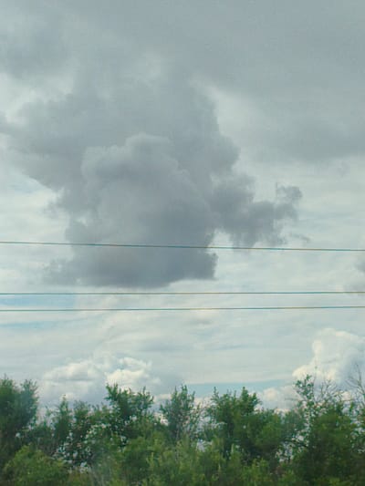 The image depicts a cloudy sky above a dense line of trees. Three power lines are visible, stretched horizontally across the frame. The sky is filled with large, grey clouds, with a slightly lighter patch of sky visible at the bottom. The trees form a dark green band at the bottom of the image, with some lighter foliage interspersed. The overall impression is of an overcast day. The location is specified as Abganerovo, Russia.