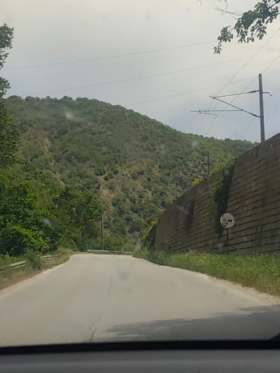 The image shows a road in Jeleznitsa, Bulgaria. The road is paved and leads uphill. On the right side of the road is a high stone wall with some vegetation growing on it. A road sign is attached to the wall, depicting a car and a person. Beyond the wall, there is green grass. On the left side of the road is more dense vegetation. In the background, a steep hillside is covered in trees. Overhead power lines run across the scene. The weather appears overcast and cloudy. The image was taken from within a car, as the dashboard is visible at the bottom of the frame.