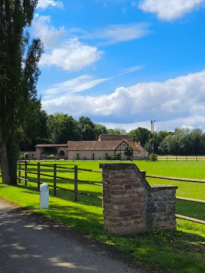The image captures a static, sunlit outdoor scene in Oberschaeffolsheim, France, under a blue sky with scattered white cumulus clouds. The foreground consists of a dark asphalt path or road on the lower left, adjacent to a tall, slender tree trunk partially covered in ivy. To the right of the path, a low stone pillar constructed from reddish-brown, rough-hewn blocks with a flat cap is positioned. Between the path and the field beyond, a multi-rail wooden fence horizontally spans the mid-left portion of the frame. A white, cylindrical bollard with a rounded top stands on the green grass immediately behind the wooden fence.
The midground features a large expanse of vibrant green grass, which recedes towards a complex of buildings. These structures possess reddish-brown tiled roofs and light-colored walls; one prominent building exhibits exposed dark timber-framing details. An adjacent structure to the left of the main building features a large, arched entrance. Beyond the buildings, a dense line of mature green trees forms a natural backdrop. In the far right background, another segment of green field is visible, extending towards a distant treeline from which a single dark utility pole rises vertically. No human figures or active subjects are present in the scene.