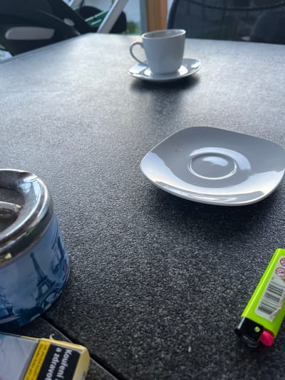The image shows a close-up of a dark gray speckled outdoor table. On the table, there is an ashtray decorated with a scene of the Eiffel Tower, a white ceramic coffee cup and saucer, and a square grey saucer. To the right of the grey saucer, a bright green lighter with a pink button is visible. In the foreground, a portion of a pack of cigarettes, with text in a yellow and black design, is present. In the background, out of focus, are parts of chairs and possibly a stroller, suggesting an outdoor seating area, such as a cafe or restaurant patio in Milná, Czechia. The lighting suggests it is daytime.