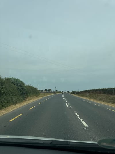 The image shows a view from a car driving on a road in Killcommon, Ireland. The road has a grey asphalt surface with yellow dashed lines on the left side and white dashed lines and solid white lines in the center. To the left of the road, there is a dense green hedge. To the right, there is a grassy area with a line of trees and a wooden fence, followed by more vegetation. The sky is overcast and grey. In the distance, there are a few cars on the road. The foreground shows the dashboard of the car.