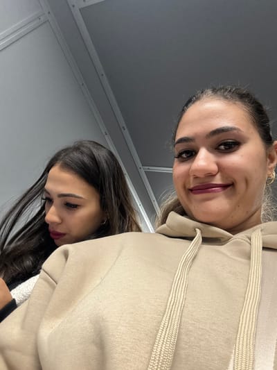 The image depicts two females in a close-up, indoor setting. The background consists of plain gray paneling with visible vertical seams and fasteners, suggesting an interior wall or partition. There are no discernible features to specify the exact location within Nouaceur, Morocco.

In the foreground, on the right, a female with a light-brown complexion and dark hair pulled back is visible from the upper torso upwards. She is wearing a light beige hooded sweatshirt with prominent drawstrings. Her face is turned towards the viewer, showing a slight smile. She has dark, arched eyebrows, extended eyelashes, and dark red lipstick. A gold-colored hoop earring is visible in her left ear.

Behind and to the left of the first female, a second female is partially visible from the chest upwards. She has dark, long hair flowing forward and a light-brown complexion. She is wearing dark red lipstick, similar to the first female. Her head is turned slightly to her right, and her gaze is directed downwards, away from the camera. She appears to be wearing a dark garment under a textured, possibly white or cream-colored, outer layer over her left shoulder. No direct interaction with the camera is observed from the second female. The image appears to be a casual photograph taken at close range.