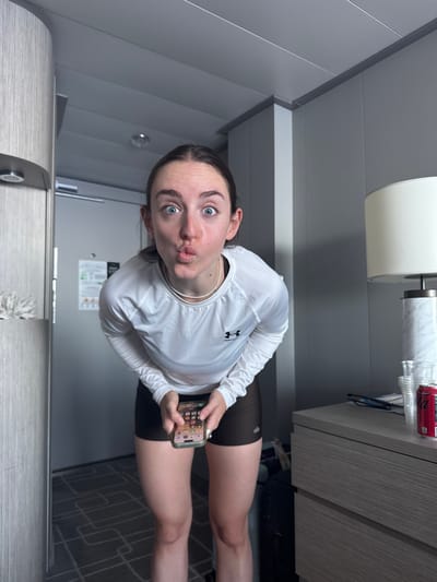 A young adult female is captured in a modern interior room, consistent with a cruise ship cabin or contemporary hotel room. She faces the viewer, leaning forward with wide blue eyes and pursed lips, holding a light-colored smartphone displaying app icons with both hands low between her legs. She wears a white long-sleeved athletic shirt with a black Under Armour logo on the chest, dark athletic shorts, and a thin, light-colored necklace. Her dark hair is pulled back.
To the left, a light-toned wooden or laminate-paneled wall unit with an integrated circular light fixture is partially visible, adjacent to a light-gray door bearing a white informational sign. To the right, a light-gray paneled wall frames a two-drawer nightstand or dresser with a light wood-grain finish. Upon the dresser are a white table lamp with a cylindrical base and fabric shade, several clear plastic cups, a pair of glasses, and a red Coca-Cola can. The floor is carpeted with a gray geometric pattern. The ceiling is composed of light-gray panels with visible seams and a small, circular light fixture. The specific location in the city of Long Island, Bahamas, is not ascertainable from the visual details within the image.