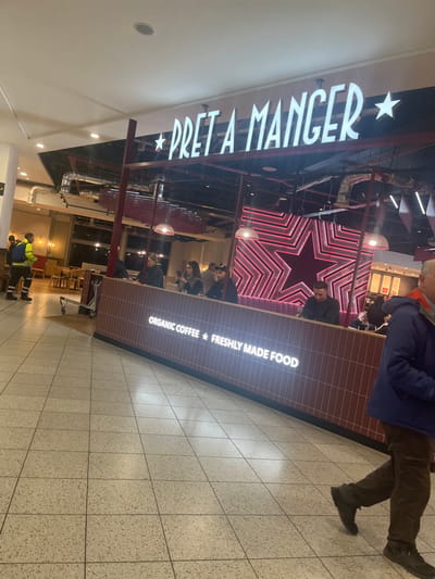 This interior photograph captures a Pret A Manger coffee shop, likely situated within a large public building such as an airport terminal or shopping center, aligning with the Horley, United Kingdom context. The perspective is from a tiled walkway, looking towards the cafe.
The prominent feature is the Pret A Manger establishment itself. A large, illuminated sign above the counter clearly displays "PRET A MANGER," flanked by star symbols. The counter area is covered in dark reddish-brown tiles, with additional illuminated text that reads "ORGANIC COFFEE * FRESHLY MADE FOOD." Behind the counter, a striking decorative element is a large, glowing red star outline, adding a vibrant touch to the cafe's interior.
Several people are visible in the scene. In the right foreground, a person wearing a dark blue jacket and brown trousers is walking past the cafe. Inside, beyond the counter, individuals are seated at tables, while one person appears to be working behind the service area. In the lower left background, two people, one of whom is wearing a yellow high-visibility vest, are also present.
The flooring consists of light-colored, square tiles, indicating a clean, public indoor space. The ceiling is high, featuring recessed lighting and visible industrial-style ductwork. The overall impression is of an active commercial environment, likely during daytime or early evening hours.