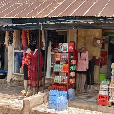 The image shows a small shop front in Kufang, Nigeria. The shop is located at latitude 9.863 and longitude 8.867, and is comprised of a corrugated metal awning and a wooden front. Clothing is hung on display from the awning, and inside the shop, several mannequins are also dressed in clothing. There is a small shelf on the right side of the shop, stocked with soft drinks and bottled water. The shop is located in a busy, bustling commercial district.