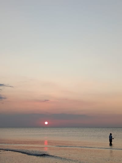 The image depicts a serene seascape at dusk. The sun, a bright pink orb, is setting on the horizon, casting a warm glow across the water and sky. Wisps of clouds, tinged with pink and orange hues, are scattered across the sky, indicating fair weather.
In the foreground, a person stands in shallow water, silhouetted against the setting sun. They are holding a fishing rod, suggesting they are engaged in the activity of fishing. The calm sea reflects the colors of the sunset, creating a mirror-like effect on the water's surface. Gentle waves lap at the shore, further enhancing the tranquil atmosphere.
The overall mood of the image is peaceful and contemplative, evoking a sense of calm and solitude. The scene is set in Jangkar, Indonesia, and the time of day is clearly sunset, characterized by the low angle of the sun and the soft, diffused light. No text is visible in the image.
