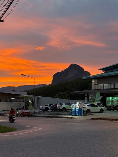 The image depicts a street scene at dusk in Ban Na Tin, Thailand. The sky is a vibrant gradient of orange and pink, with a dark, conical mountain visible in the background, silhouetted against the colorful sky. In the foreground, a street lamp with a red-lit head stands on the right side of the road. A road with a white lane marking curves through the scene. On the right side of the road, there is a building with a green awning and large windows, with a sign that reads "NOW OPEN!" and some Thai script. Adjacent to the building is a gas station with blue pumps and a white car. Several other cars are parked in the background, including a white pickup truck. On the left side of the road, a person on a scooter is riding past. The overall environment suggests an urban or semi-urban setting within a mountainous region.
