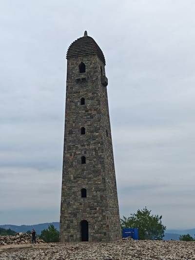 This is a full-frame, eye-level, outdoor, medium shot photograph of the medieval "tower of the dead" in the village of Эникали, Russia. The tower, constructed of rough-cut stone, is tall and slender, tapering slightly as it rises. It has a conical roof topped with a pointed spire. Numerous small, arched windows are visible at various levels of the tower. The tower is surrounded by a rocky, uneven terrain. In the foreground, a man is standing on the rocks, facing the tower and holding a camera, appearing to take a photograph. To the left of the man, a large pile of stones is visible. Behind the tower, green trees and rolling hills can be seen against a cloudy, overcast sky. A blue tarp covering some construction materials is partially visible to the right of the tower. The overall scene depicts a historical monument in a somewhat rural and undeveloped setting.