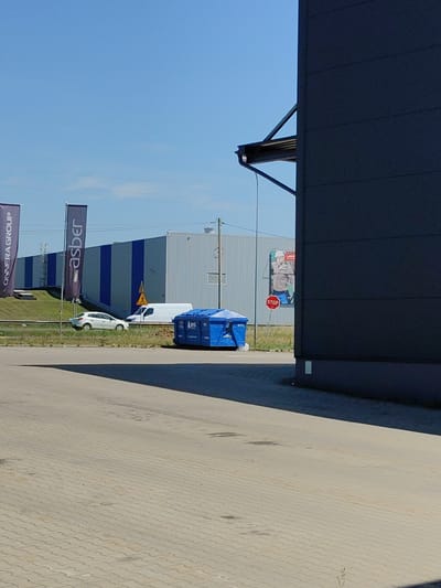 In Łomna-Las, Poland, a bright, sunny day illuminates a paved area in front of a modern building with a dark gray facade. Two tall, dark banners stand on the left, one displaying "ONNERA GROUP" vertically and the other with "asber". Adjacent to these banners is a white van and a white car parked on a grassy verge. Further into the scene, a large blue waste bin is situated. Behind the vehicles, a long industrial building with alternating blue and white vertical stripes is visible. A billboard featuring a man in workwear stands on the right side of the industrial building, accompanied by a red "STOP" sign. The sky is clear and blue, with a few power lines visible in the distance.