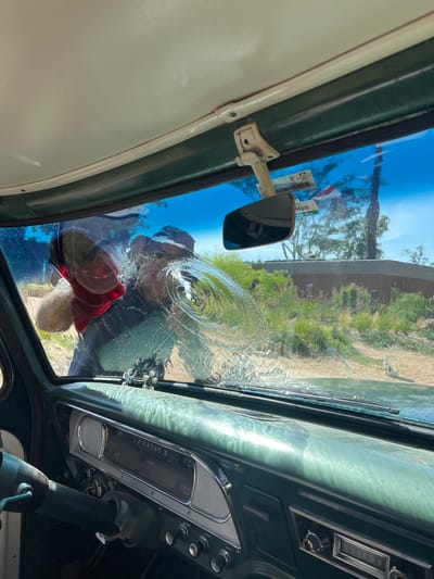 The scene is observed from within the cabin of an older model vehicle, looking outward through the windshield. The vehicle's interior features a dark green dashboard with a silver-colored instrument panel bezel and a partially visible dark green steering wheel on the left. A small, dark decorative object resembling a miniature guitar is positioned on the dashboard near the windshield. A rearview mirror is attached to the light-colored headliner.
Outside the vehicle, a person is actively engaged in cleaning the windshield. The individual is partially obscured and distorted by the water on the glass, appearing to wear dark clothing and a hat. A red glove or cloth is visible on their right hand, making contact with the windshield. Water is visibly present across the glass, forming concentric circular patterns and streaking downwards.
The exterior environment indicates a sunny day with a clear blue sky. The background features light-colored ground, possibly dirt or sand, with sparse green vegetation, including shrubs and several trees. A low, dark wall or structure is visible further in the background, and a white, rectangular object on a pole stands to the right. Vehicle identification stickers, one displaying the number "98509," are affixed to the upper right area of the windshield.
This scene is located in La Barra, Uruguay.