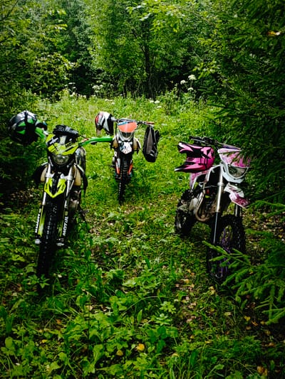 Three dirt bikes are parked in a wooded area, surrounded by lush green foliage and trees. The bikes are positioned in a diagonal line from left to right.
The leftmost bike is bright green and yellow, with a black and green helmet resting on its handlebars. The middle bike is orange and white, with a red, white, and black helmet on its handlebars. A black backpack is also hanging from its handlebars. The rightmost bike is white and pink, with a matching pink helmet on its handlebars. The bikes are all facing forward.
The ground is covered in grass and fallen leaves. The trees in the background are dense and green, indicating a temperate climate. The overall scene suggests an outdoor recreational activity.
The location of Rassol'naya, Russia is not explicitly visible or deducible from the image itself.
