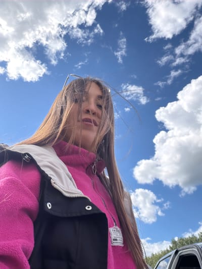 A low-angle, medium shot captures a young woman with long, brown hair against a bright blue sky dotted with white clouds. She is wearing a hot pink fleece jacket layered under a black vest with a tan collar. Her hair is windblown, partially obscuring her face. The background shows the tops of green trees and a portion of a car. The scene appears to be outdoors in an open area.