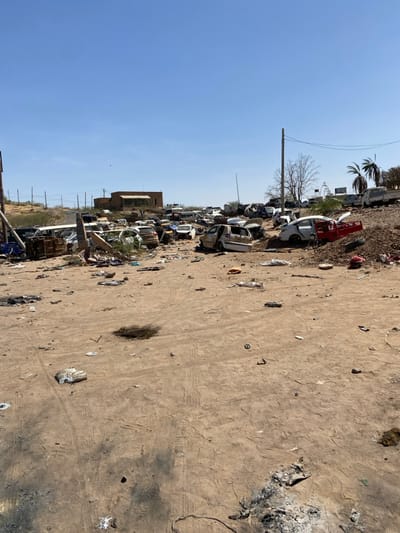 The image depicts a scene of destruction and disarray in Tayba Alhasanab, Sudan. It is a sandy, open area, strewn with debris and wreckage of multiple vehicles. The vehicles are mostly overturned and damaged. There are no people visible in the image. The scene is likely the aftermath of an incident or conflict. A building is visible in the background. The sky is clear and blue.