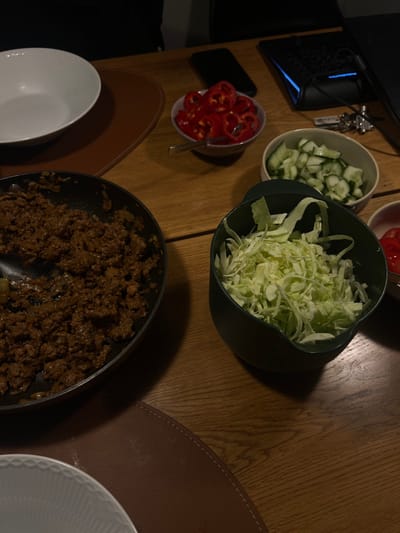 The image shows a wooden table with various ingredients for a meal. The ingredients include a large skillet filled with cooked ground meat, a bowl of chopped cabbage, a bowl of chopped cucumbers, and a bowl of chopped red bell peppers. There is a white plate on the left side of the image and a brown place mat on the right side. The table is located in a home kitchen in the city of Skanderborg, Denmark. The location is identified by coordinates (latitude: 56.049, longitude: 9.926).