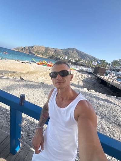 In the city of Pla Parcial Platja, Spain, a man is taking a selfie on a beach. He is wearing a white tank top and black sunglasses. The beach is covered in pebbles, and there are colorful paddle boats lined up on the shore. In the background, there are mountains, palm trees, and buildings. The sky is clear and blue. A can of soda is on a blue railing in the foreground.