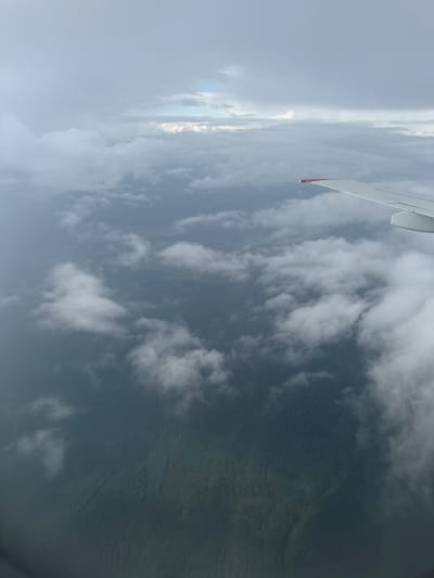 The image is an aerial view of a densely forested landscape, likely taken from an airplane. The terrain appears to be characterized by rolling hills covered in thick, dark green vegetation, indicative of a forest. Patches of clouds are scattered across the scene, obscuring parts of the forest below and creating varying degrees of light and shadow. In the upper right portion of the image, a portion of an airplane wing is visible, featuring a red tip, suggesting the perspective is from inside an aircraft. The sky above is overcast with a mixture of light and dark gray clouds, with a sliver of brighter sky peeking through in the distance. No human presence or specific structures are discernible within the visible forest or landscape. The location in the city of Churr, Russia is not identifiable from the visual information in the image alone, as it depicts a natural, undeveloped environment.