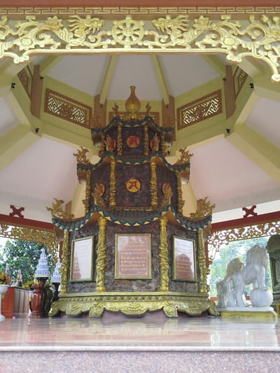 The image depicts an ornate, tiered structure within a pavilion in Thiện Chí, Vietnam. The structure, featuring intricate carvings and religious symbols, is adorned with gold accents and topped with a decorative urn. It is flanked by framed inscriptions and seated on a base that resembles rough stonework, painted gold. A pair of white lion statues, one with a paw resting on a sphere, stands to the right of the structure. The pavilion has an open design, revealing a view of lush greenery outside. The interior features a decorated ceiling with geometric patterns and golden ornamentation, and the floor is highly polished, reflecting the surrounding elements. The scene suggests a religious or cultural setting.