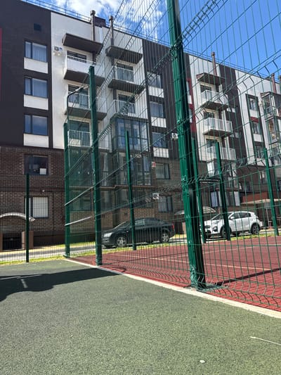 This is a vertical, eye-level shot of an apartment building and a sports field, taken on a sunny day in Viazniky, Russia. The building is tall and modern, with a dark brown and white facade, featuring numerous windows and balconies. A green metal fence runs across the foreground, partially obscuring the view of the building and a sports field with a red running track and a green artificial turf area. Two cars are visible parked in front of the building, one white and one dark. The sky is blue with scattered white clouds. The overall impression is one of an urban residential area with recreational facilities. There is no visible text in the image.