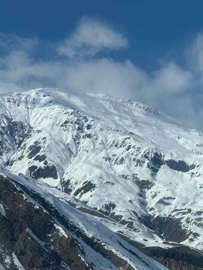 The image presents a snow-covered mountain landscape under a blue sky with scattered white clouds, located in Kanobi, Georgia. The primary subject is a large mountain, extensively covered in white snow that highlights its complex topography of ridges, valleys, and slopes. Numerous dark, jagged rock outcrops are visible through the snow, particularly on the steeper sections and lower-lying areas. The snow exhibits varied textures and depths, casting shadows that define the mountain's contours. The sky occupies the upper portion of the frame, featuring a clear blue expanse interspersed with white, fluffy cumulus clouds. Some thin, wispy cloud formations are present near the mountain peaks. No individuals, human-made structures, or actions are depicted within the scene. The environment is characteristic of a high-altitude, winter mountainous region.
