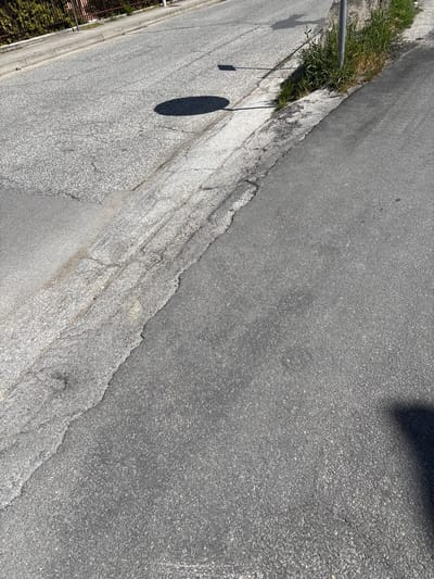 This image captures a ground-level view of an outdoor urban environment, likely a street or paved area in Camerino, Italy, under bright sunlight. The scene is dominated by asphalt surfaces, displaying a clear contrast between a darker, seemingly newer or smoother section in the upper right and a lighter, older, and more worn section that occupies the majority of the left and central frame.

The lighter asphalt exhibits numerous cracks and a rougher texture, indicative of wear and tear, and features an irregular, jagged boundary with the darker asphalt. Along the far left side, a concrete curb or edge borders the lighter road surface. Adjacent to this curb is a strip of unkempt green grass and weeds, suggesting natural growth in the urban setting. A thin, grey metal pole, possibly a signpost or utility pole, is visible emerging from this grassy strip near the top left.

Several shadows are cast across the surfaces, indicating a clear, sunny day, likely mid-morning to late afternoon. A distinct, dark, oblong shadow lies across the lighter asphalt in the mid-left, while a thin, linear shadow extends from the metal pole. A larger, irregular dark shadow is also visible in the upper right corner of the frame, partially covering the darker asphalt. In the very bottom left, a dark metal fence or railing is faintly visible behind some green foliage. There are no people or specific activities depicted, and no visible text is present in the image.