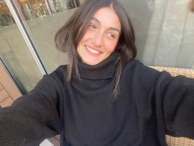 A light-skinned female individual, appearing to be a young adult, is captured in a close-up, self-portrait style photograph. She has dark brown, shoulder-length hair and is smiling broadly, with her head slightly tilted, gazing towards the viewer's upper left. She is wearing a dark, likely black, knit turtleneck sweater, which shows visible light-colored lint or fibers, particularly on the sleeves. Her left arm, extended to hold the camera, is partially visible in the lower right of the frame. The background on the left consists of a large glass window reflecting an indistinct interior, possibly a retail space, with a light-colored wooden panel below it. On the right, a section of a light-brown wicker or woven chair back is visible, indicating an outdoor or semi-outdoor seating environment. The scene is brightly illuminated by natural light, suggesting a daytime setting within a commercial or promenade area in the city of La Grande-Motte, France.