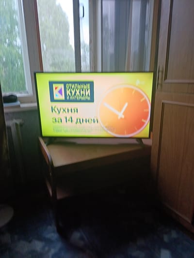 The image shows a television screen displaying an advertisement for "Stylish Kitchens and Interiors" with the slogan "Kitchen in 14 days". The advertisement features a logo with a stylized "K" and a clock face with orange and white colors. The TV is placed on a wooden stand in front of a window with vertical blinds and curtains. The overall scene appears to be an indoor setting, possibly a living room or bedroom, in Koryazhma, Russia. The lighting suggests it might be daytime, with natural light coming from the window. The image is slightly blurry, indicating a handheld shot or low light conditions.