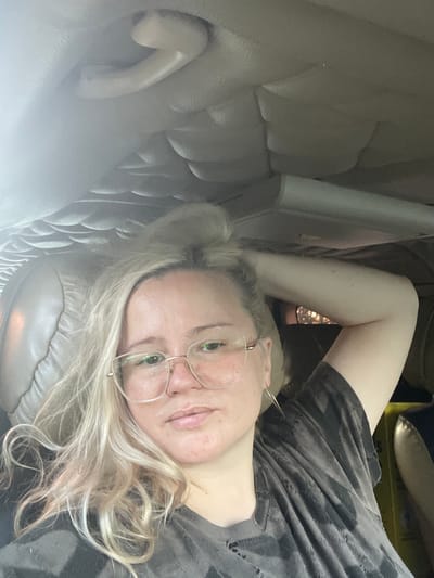 This media file shows a woman lying down in a car. The woman has blonde hair and is wearing glasses and a dark, patterned t-shirt. She is looking up towards the camera, and her expression appears relaxed. The interior of the car is visible, with a light-colored leather seat. The lighting suggests it is daytime, possibly late afternoon or early evening, as there is a warm glow coming from outside. The image is taken from a low angle, looking up at the woman. There is no visible text in the image.