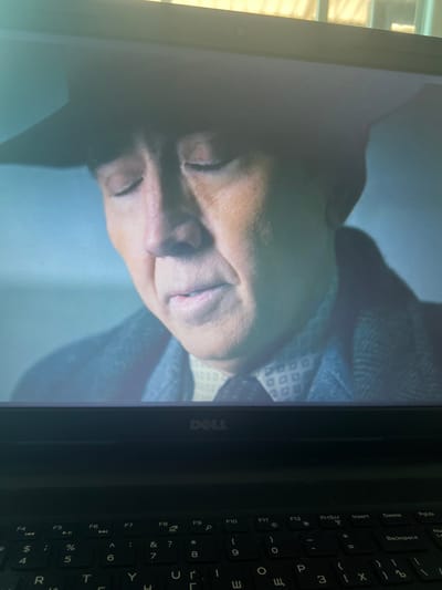 The image shows a close-up of a person's face from the chest up, displayed on a laptop screen. The person appears to be wearing a hat and a patterned shirt under a dark jacket. The lighting suggests an indoor setting, possibly a room with ambient light from a window. The lower portion of the image shows a black Dell laptop keyboard, with keys like F4, F5, F6, F7, F8, F9, F10, F11, F12, PrtScn, Insert, Delete, and PgUp visible. The scene is not specifically located in Nikopol, Ukraine, as no identifiable landmarks or context are provided.