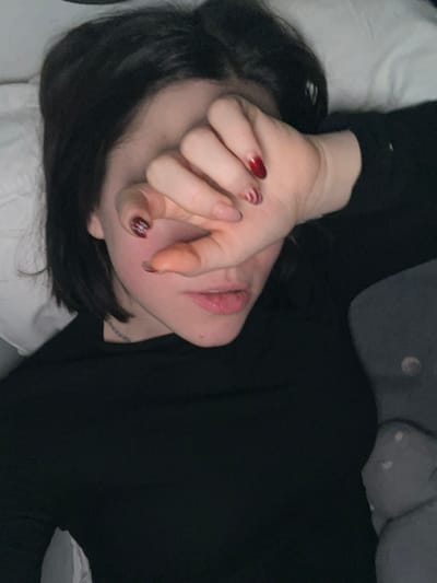 A person lies in bed wearing a black top. They have short dark hair and their face is partially obscured by their hand. The hand has manicured nails, some with red polish and some with a white swirl design. The person's lips are visible and slightly parted. Part of a white pillow is beneath their head, and a grey patterned pillow is visible in the upper portion of the image.