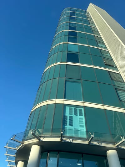 A tall, modern building with a curved glass facade is depicted against a clear, bright blue sky in Gillingham, United Kingdom. The structure's primary face consists of multiple stories of teal-tinted glass panels framed by light-colored horizontal bands and dark vertical mullions. To the right, a contrasting, flat vertical section of the building rises, clad in light-colored, possibly ribbed, panels. At the base of the curved section, a cantilevered glass balcony or awning extends, supported by light-colored cylindrical columns and metal brackets. One window on a lower floor of the curved section clearly displays closed white louvered shutters, while an upper window on the flat section appears to be slightly ajar. Strong sunlight illuminates the scene, casting distinct shadows from the balcony supports onto the building's facade. No individuals, actions, or specific interactions are visible.