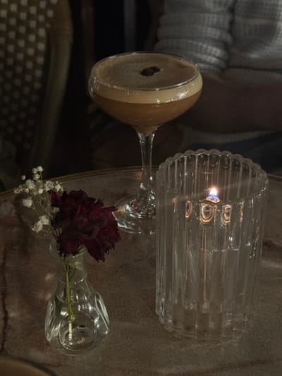 A martini glass with a coffee-colored drink rests alongside a ribbed glass candle holder with a lit candle. Nearby, a small glass vase holds a deep red carnation and sprigs of white baby's breath. All items sit on a textured surface. A blurry light-colored sweater is in the background.