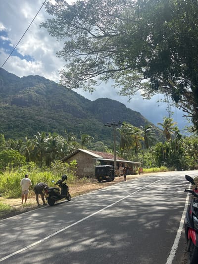 This is a rotated, eye-level, outdoor shot showing a roadside scene in Moneragala, Sri Lanka. The time of day appears to be late morning or early afternoon, judging by the strong shadows cast by the trees on the road. The sky is partly cloudy, with areas of bright sunshine and darker, more dramatic cloud formations.
The scene is dominated by a lush, green mountain covered in dense vegetation and dotted with palm trees in the mid-ground. To the right of the mountain, a narrow asphalt road runs diagonally through the frame, marked with a white line. The road is shaded by dappled sunlight filtering through the leaves of trees on the left.
Along the side of the road, there's a small, rustic building with a tiled roof and brick walls. In front of this building are a few small stalls, possibly selling goods. A dark-colored auto-rickshaw is parked nearby.
Several people are present. Two individuals, dressed in casual attire, are interacting with a parked black scooter on the side of the road. Another person in a light-colored shirt stands further down the road, facing away from the camera. The overall atmosphere seems relaxed and unhurried. The dense foliage, palm trees, and the style of the building and vehicles suggest a tropical or subtropical environment. No visible text is discernible.