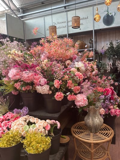 This is an eye-level, vertical, medium shot of a vibrant display of artificial flowers in what appears to be a store or market in Tuk, Netherlands. The subjects are an abundance of various artificial flowers in shades of pink, purple, yellow, and white. The flowers are arranged in large groupings, some in dark buckets or containers, creating a lush and abundant visual.
Notable objects include several large, dark, cylindrical buckets holding the flowers, a woven rattan chair with a circular design, and a tall, decorative vase with a marbled gray and white pattern placed on the chair. Hanging from the ceiling, illuminated by light bulbs, are at least two pendant lamps with woven shades, and another large, Edison-style bulb hangs by a wire.
The setting appears to be indoors, possibly a greenhouse or flower shop, with a wooden plank ceiling and exposed metal beams of a larger structure above. There are also indications of shelving or displays in the background.
Visible text includes "Ja, bloemen voer" and "materiaal van" on a light-colored panel in the background, which seems to be promotional material for the flowers. There are also small white tags on some of the flower stems, typical of retail items.
The overall impression is one of a colorful and abundant floral display, likely aimed at attracting customers. The lighting suggests it could be daytime, though the exact time of day is not discernible. There are no people or animals visible, and the focus is entirely on the floral arrangement and associated decorative objects.