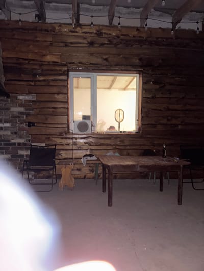 This is a medium-shot, eye-level photograph taken at night, depicting an outdoor covered patio area. The patio features a wooden plank wall with a large window. Inside the window, there is a fan and some objects on a shelf, illuminated by indoor lighting. In front of the wall, there is a wooden table with a bottle of beer and some other items. To the left of the table, there is a black folding chair and a wooden stool. Further to the left, a brick wall is visible. The ground is concrete. The overall atmosphere is rustic and casual. The image's lighting is low, suggesting it was taken in the evening or at night. The scene does not contain any identifiable landmarks of the city of Saran, Kazakhstan.