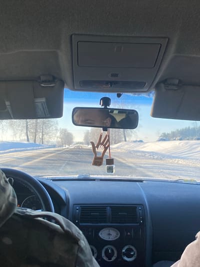 The image depicts the interior of a vehicle from a front passenger perspective, looking forward through the windshield onto a snowy rural road in Карабоўскі сельскі Савет, Belarus. The exterior environment shows a two-lane asphalt road, partially cleared of snow, stretching into the distance under a bright, clear sky. Deep snowbanks line both sides of the road, and bare deciduous trees, appearing white with frost or snow, are visible in the middle distance.
Inside the vehicle, the dark-colored dashboard and a black multi-function steering wheel are prominent. The driver's left shoulder and the back of their head are partially visible on the left, clad in a camouflage-patterned jacket. The central rearview mirror reflects the driver's face, showing a male individual with short hair. Suspended from the rearview mirror are two items: a light-brown wooden ornament shaped like an open hand performing a Vulcan salute, with Cyrillic text "Жыве Беларусь!" (Long live Belarus!) inscribed on it, and a small, square glass bottle containing a Lacoste-branded air freshener. The vehicle's light-colored fabric headliner, an overhead console with a rectangular compartment, and both stowed sun visors are visible in the upper portion of the frame.