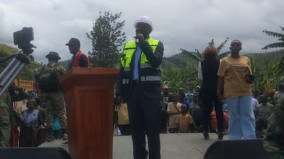 The scene is a public gathering in Sake, Democratic Republic of Congo, situated at latitude -1.57, longitude 29.046. A man wearing a white hard hat and a yellow reflective vest is speaking into a microphone at a podium. The podium is made of wood and is standing on a paved platform. He is dressed in a dark blue suit and a light blue shirt. Behind him is a large crowd of people. A woman wearing a tan t-shirt and blue jeans is standing to the right of the podium. Two men are standing in front of the podium, one wearing a military uniform and the other wearing a red vest. A video camera mounted on a tripod is positioned to the left of the podium. The background is a hilly landscape with trees and vegetation. The sky is overcast with clouds. A group of children are seated in the front of the crowd.