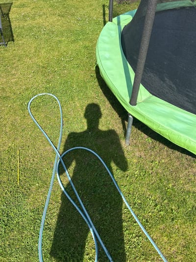 In Haaslava, Estonia, a sunny day illuminates a grassy lawn where a trampoline and a coiled garden hose are present. The trampoline, partially visible, features a black jumping mat and a bright green safety pad. A person's shadow, cast by the sun, falls across the grass, overlaying the coiled hose, which is light blue with lighter blue stripes. A single yellow marker stake is also visible in the grass. The scene appears to be a backyard setting.
