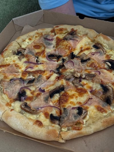 A close-up, overhead shot shows a large, round pizza with ham, mushroom, and red onion toppings in a brown cardboard pizza box. The pizza is cut into eight slices, and some pieces are slightly askew. The crust is golden brown and dusted with flour. The background is a blurry green surface, possibly a tablecloth, and a person's arm wearing a blue shirt. The scene is located in Nova Odesa, Ukraine.