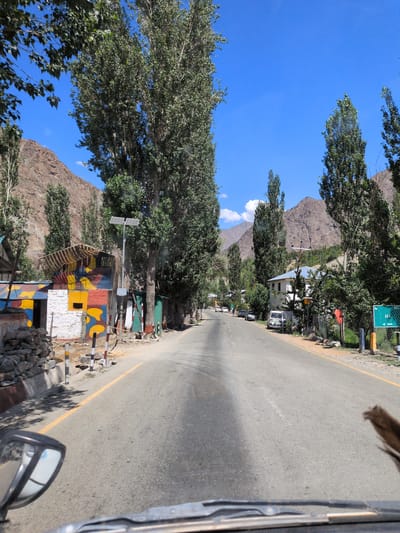 The image shows a view from inside a vehicle traveling on a paved road in a village, likely in Chanigund, India, as suggested. The road has a yellow dividing line. On the left side of the road, there is a building with brightly colored murals, and beyond that, a stone wall and trees. A tall solar-powered lamp is positioned near the building. On the right side of the road, there are more trees, houses, parked cars, and a green road sign. The surrounding environment features dry, rocky mountains and a clear blue sky. The presence of trees and a building suggest a rural or semi-urban setting.