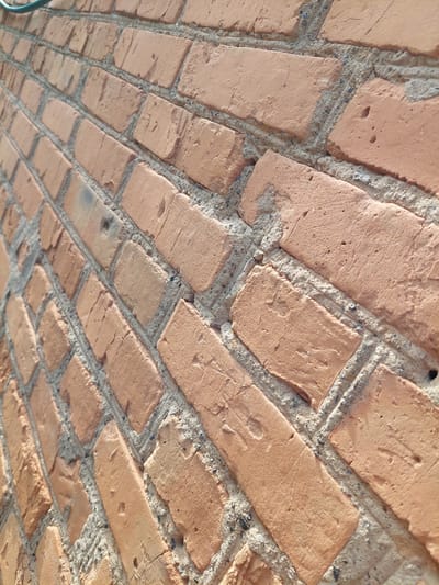 The image shows a close-up, angled view of a brick wall constructed with rough, reddish-brown bricks and grey mortar. The bricks are laid in a staggered pattern, and many show signs of wear, including cracks, chips, and small holes. The mortar also appears somewhat uneven and textured. The lighting suggests it is daytime, casting shadows that emphasize the texture of the wall. A small sliver of a green pipe or cable is visible in the upper left corner of the frame. The scene depicts a section of an outdoor wall, likely in a residential or industrial area, as no people or specific urban landmarks are identifiable. Based on the provided information, this wall is located in the city of Shpychyntsi, Ukraine.