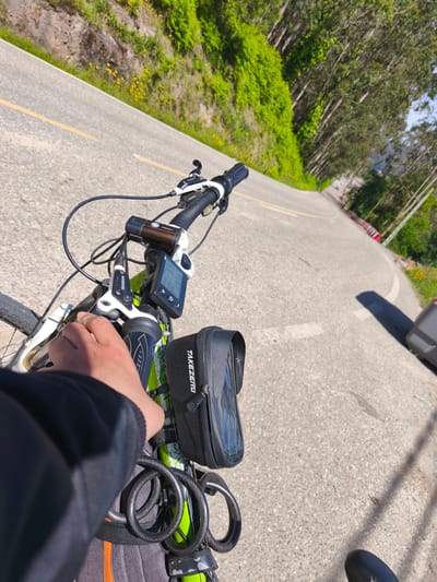 A first-person perspective shot taken from a bicycle on a curved road in Cangas de Morrazo, Spain. The foreground includes the bike's handlebars, a digital display, a "TAKEZERO" bag, a lock, and a hand gripping the brake. The road is asphalt, marked with a yellow line along the left side. A green, foliage-covered hill rises along the left, and a line of trees extends along the right side of the road. A grey car is partially visible on the right side of the road. Red and white markers line the road further ahead, suggesting a curve or turn. The sky is visible in the upper right corner.